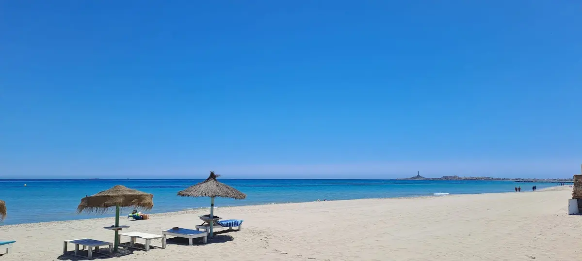 Sandy Mediterranean beach with straw parasols and the Cabo de Palos lighthouse in the distance